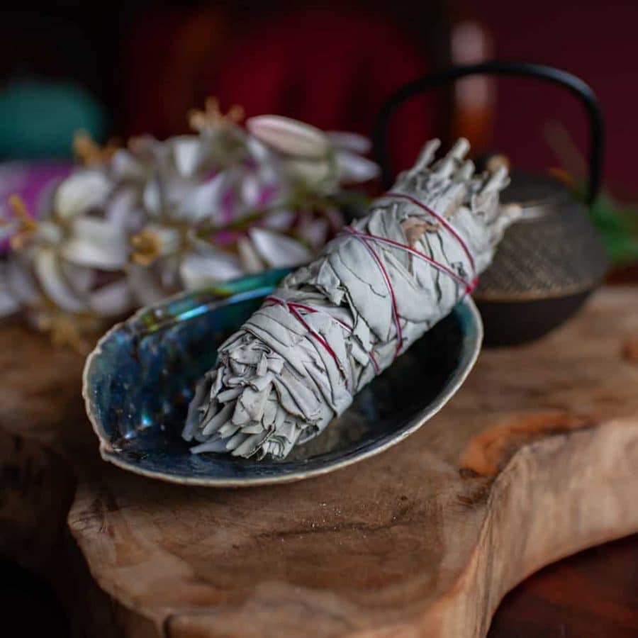 Smudge-Stick Weißer Salbei in blau schimmernder Abalonemuschel auf Holz mit Blumen und Teepott im Hintergrund