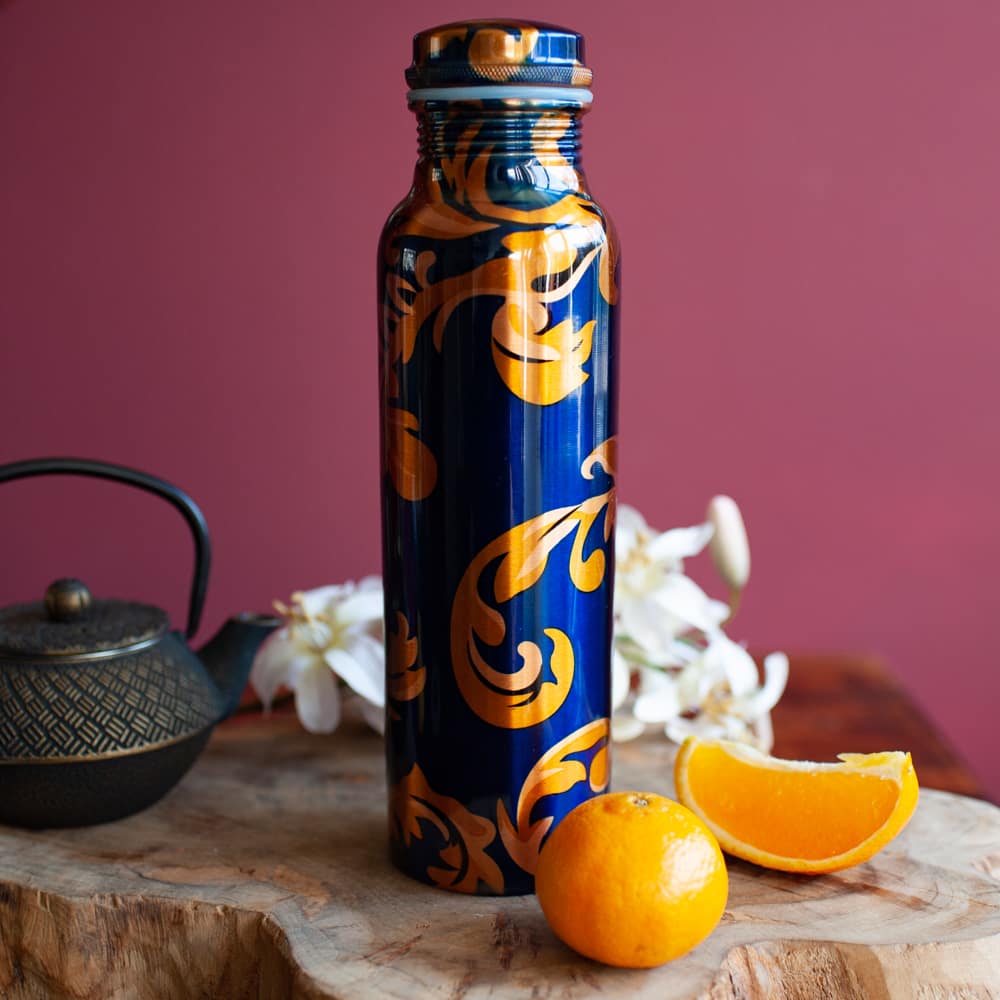 Blaue Kupferflasche mit floralem Design vor roter Wand auf Holz mit Orangen und Teepot