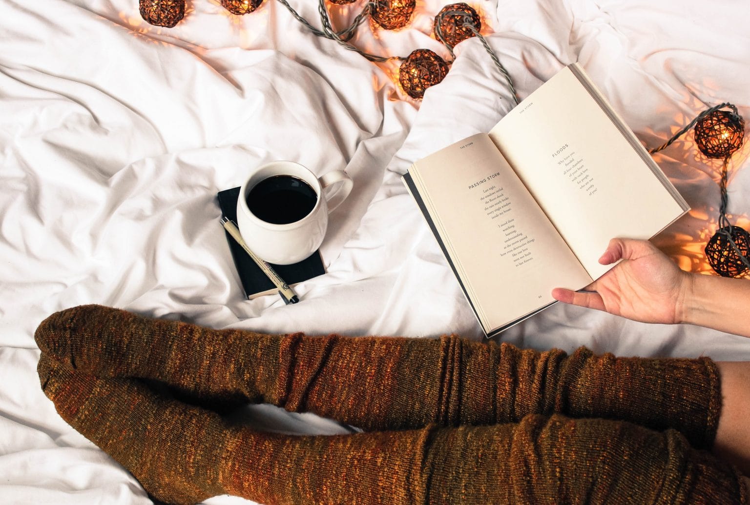 Beine mit hohen braunen Socken mit Buch und Kaffee auf weißer Bettdecke mit Lichterkette